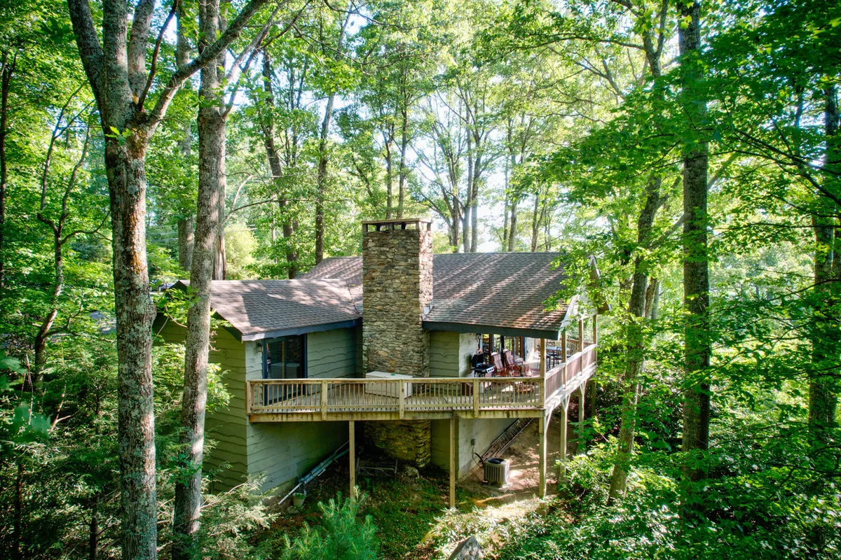 The image showcases the exterior of a beautifully renovated mountain home nestled among tall trees, featuring a striking stone chimney and an inviting wooden deck that overlooks the tranquil Shulls Mill Valley. The deck offers ample space for relaxing, enjoying nature, and gathering with loved ones—all set against a backdrop of serene forested hills.