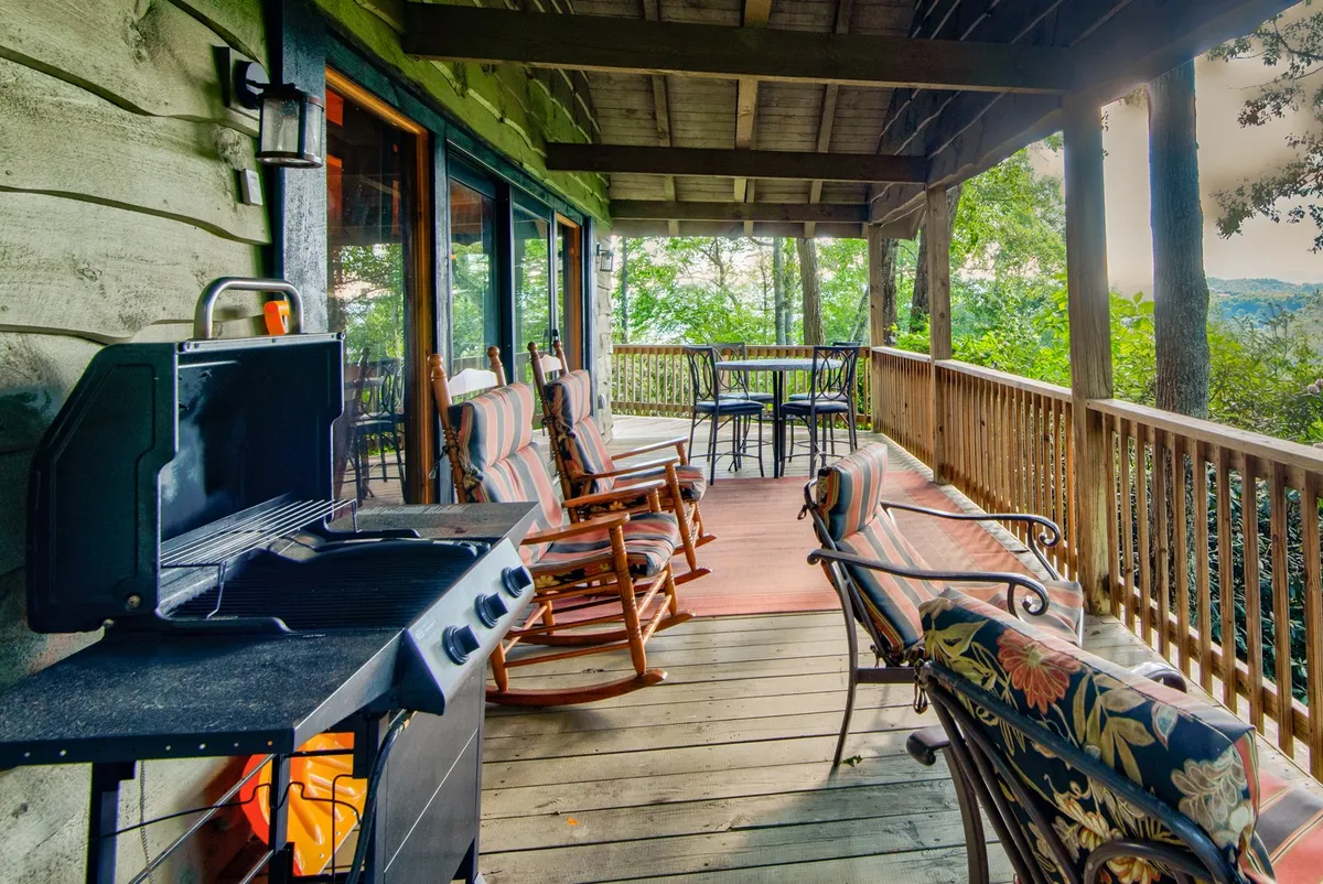 An exterior deck scene featuring a gas grill, rocking chairs, a table and chairs, and a scenic outlook of trees and rolling hills. The setting is casual and serene, ideal for dining, grilling, and enjoying the natural landscape in any season.