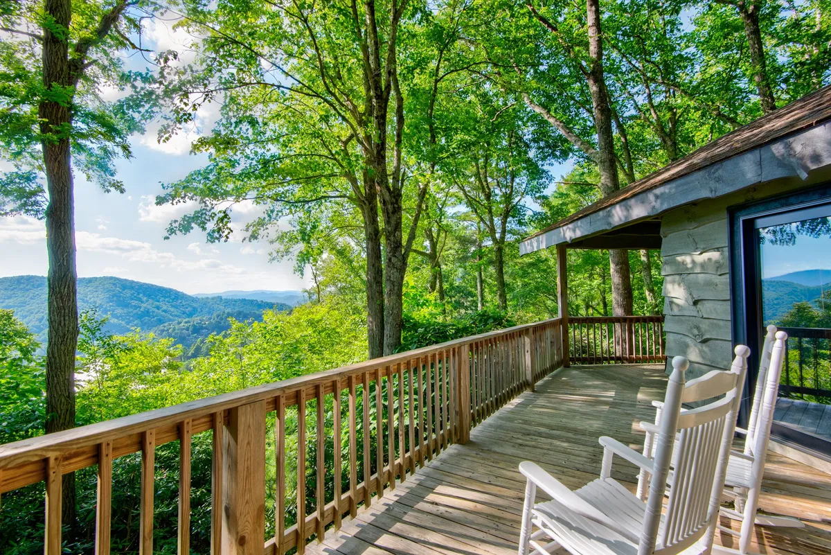 A wooden deck with classic rocking chairs and a sturdy railing frames a sweeping mountain view. Lush greenery encircles the deck, inviting guests to relax, read, or socialize while taking in the tranquil natural surroundings.