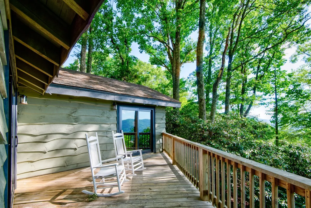 A wooden deck with rocking chairs positioned to overlook a mountain view through the trees. The mood is relaxed, with soft natural lighting that hints at either sunrise or sunset. The setup invites quiet conversation and leisurely enjoyment of the outdoors, emphasizing peaceful togetherness.