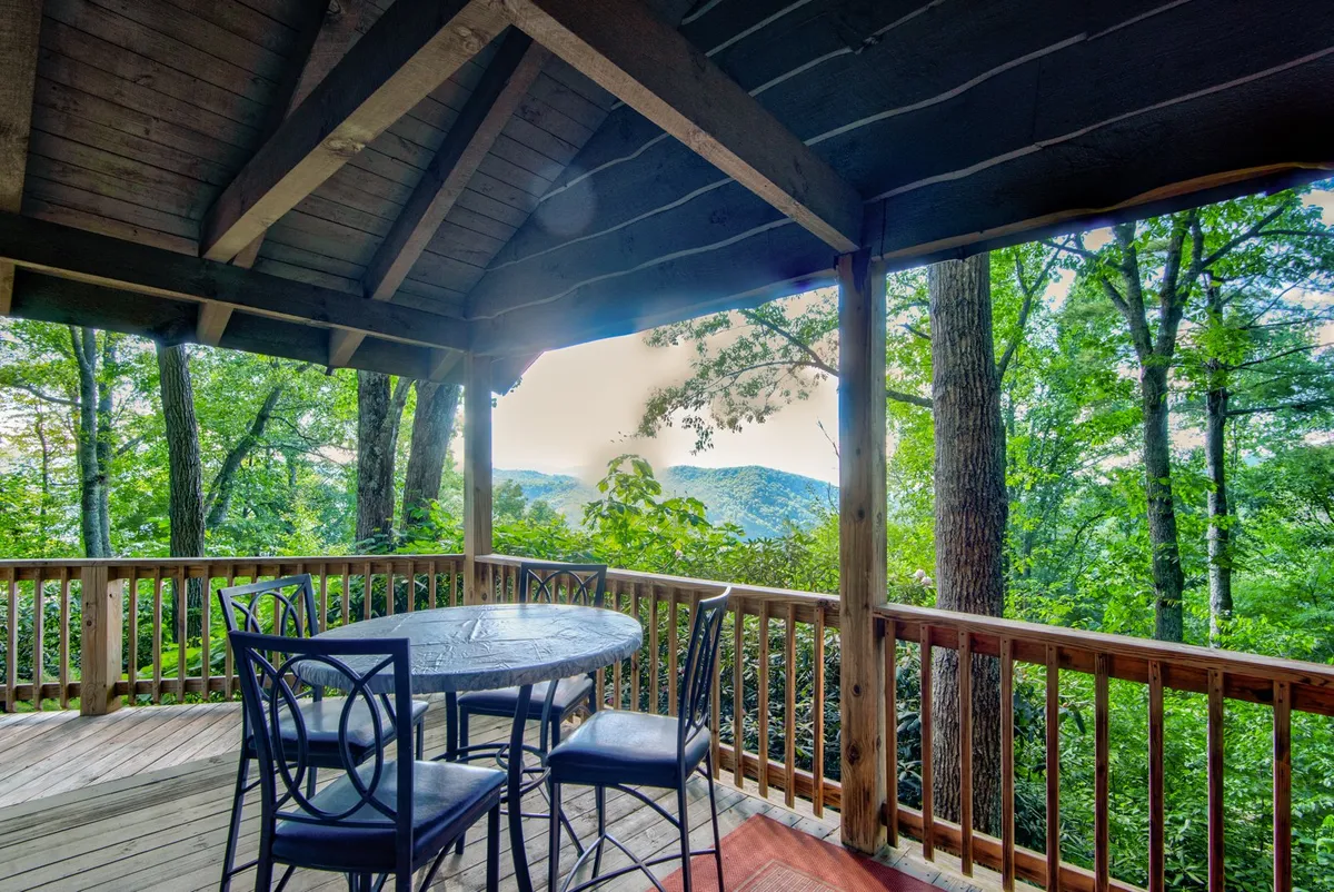 A deck space with a dining table, chairs, wooden railing, and unobstructed mountain views through the trees. The scene is peaceful, designed for enjoying meals outdoors or soaking up the picturesque surroundings.