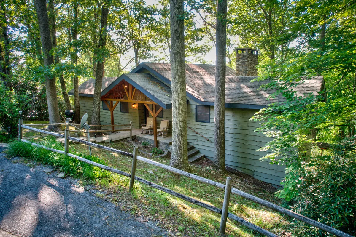 This exterior image showcases the wooden porch of Crystal View, featuring a sturdy stone chimney and lush surrounding trees. The porch appears inviting for relaxing, while the spacious outdoor area hints at opportunities for gathering, barbecuing, or enjoying nature. The tranquil, cozy mood is emphasized by the natural setting and the gathering-friendly features.