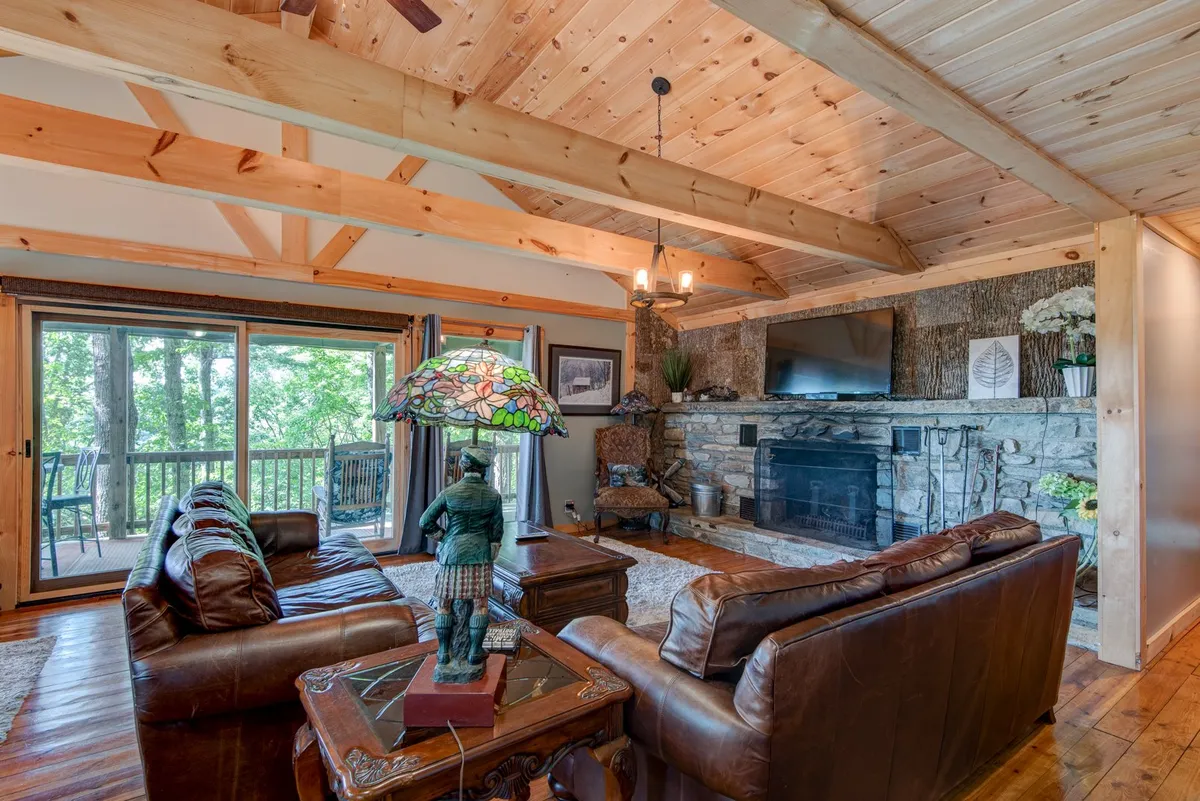 A spacious great room with soaring wooden beams and a peaked beadboard ceiling. The central feature is a large stone woodburning fireplace, flanked by leather sofas and a stained glass lamp. Large windows span the back wall, filling the room with natural light and offering tree-filled views that bring the outdoors in.