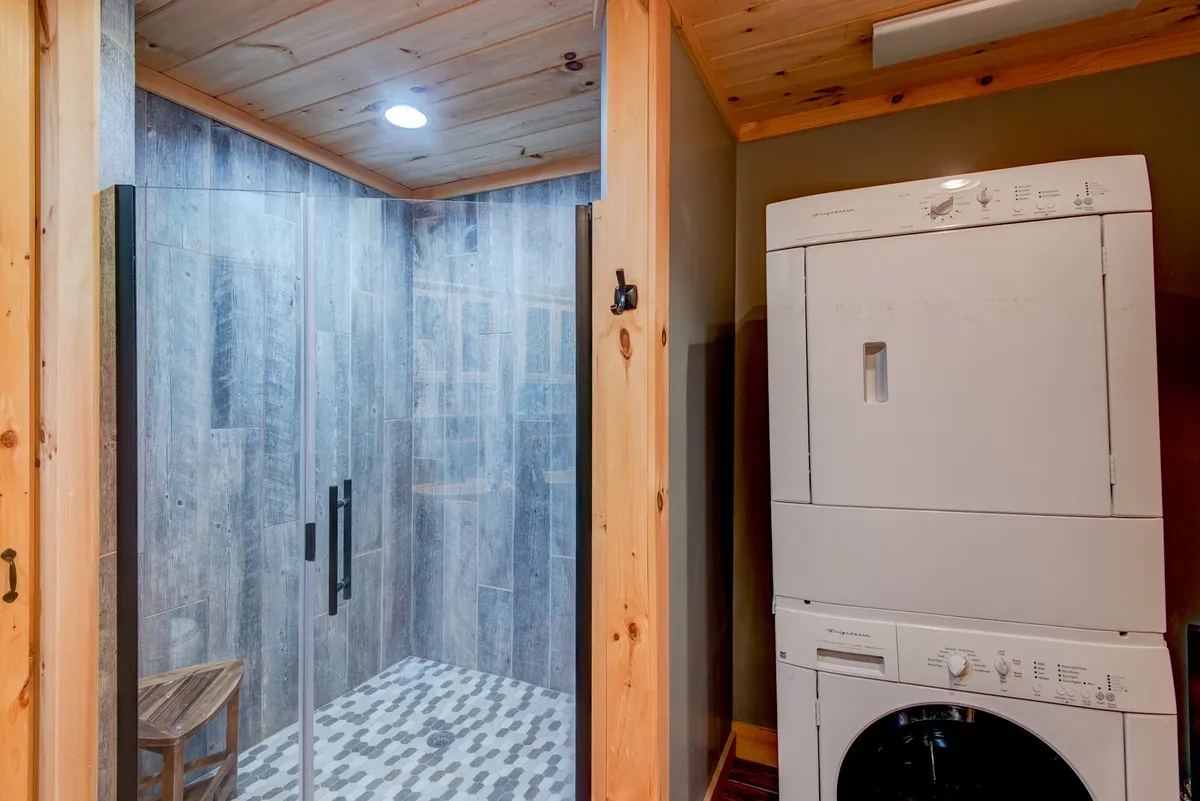 The image shows a practical yet serene space with a modern shower area, washer and dryer unit, and a wooden chair. Clean lines and natural textures create a tranquil setting for both daily routines and extended stays.