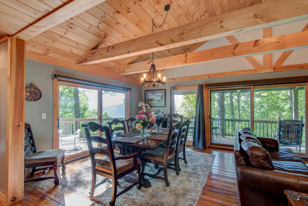 A beautifully carved dining table with a floral centerpiece is set in front of expansive windows offering a scenic view of the mountains. A statement chandelier hangs overhead, and soft natural light fills the space, enhancing the warm, welcoming mood.