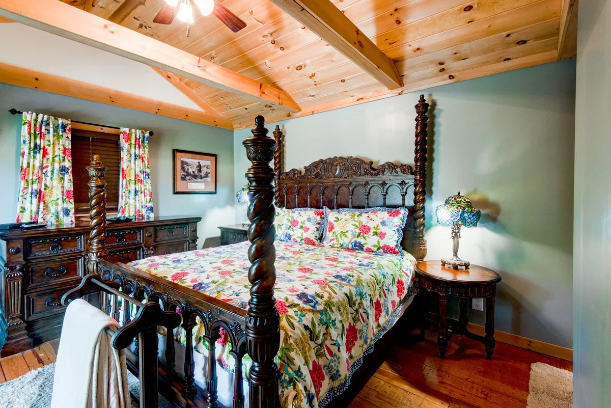 The image presents a cozy bedroom with a four-poster bed adorned in colorful floral bedding, ornate wooden furniture, a stained glass lamp, and a window with patterned curtains. The setting is warm and inviting, ideal for restful nights or a quiet morning.